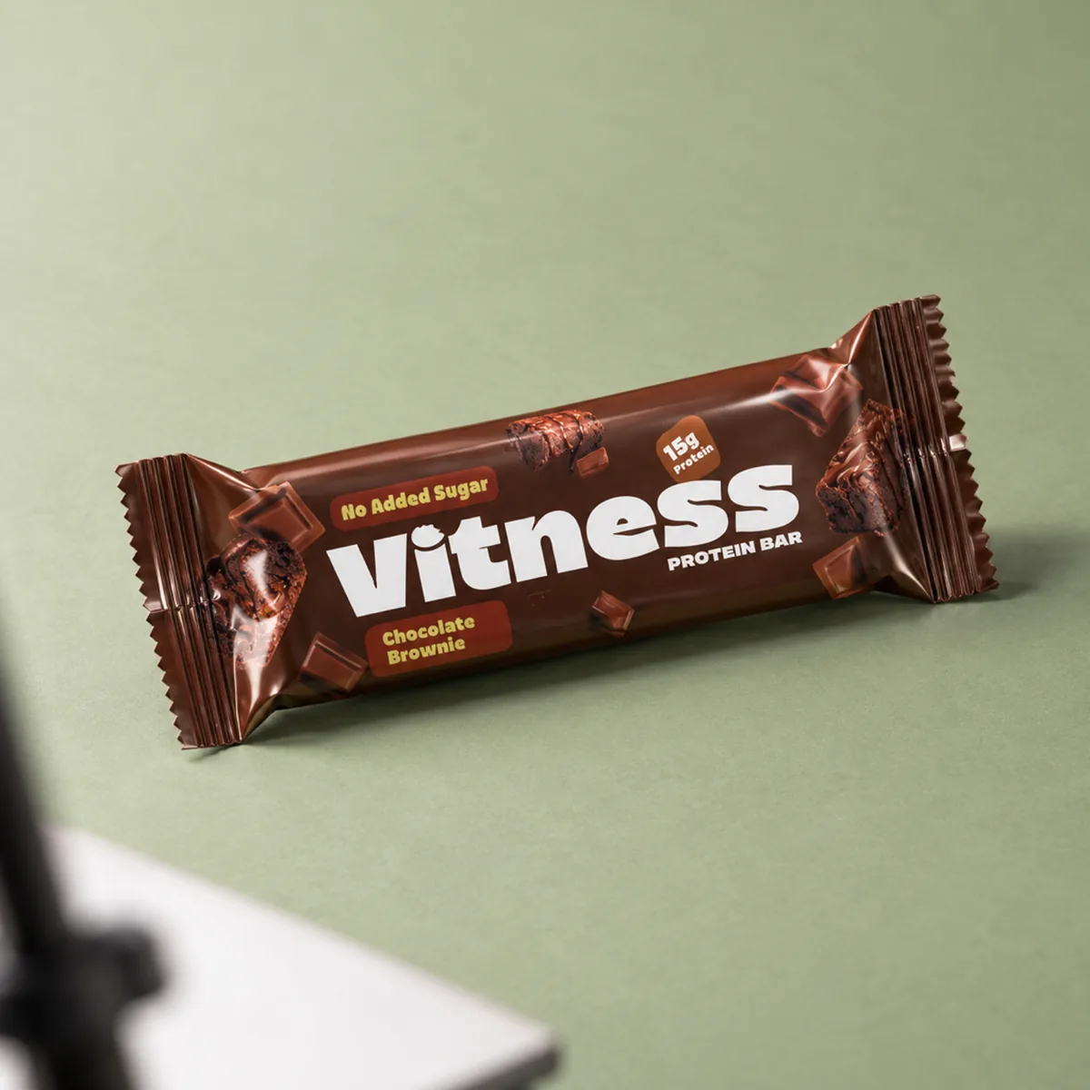 A sealed Vitness protein bar pack on a sage backdrop with a reflector card in the foreground