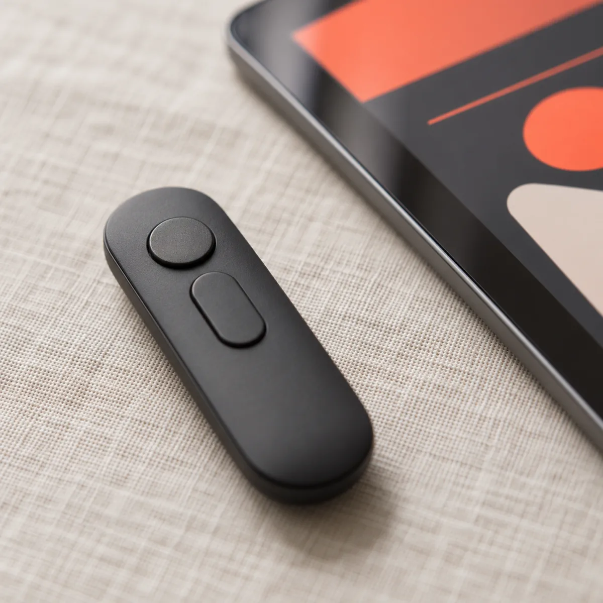 Close-up of a matte charcoal presentation clicker on linen beside a tablet showing abstract coral and charcoal slide layouts.