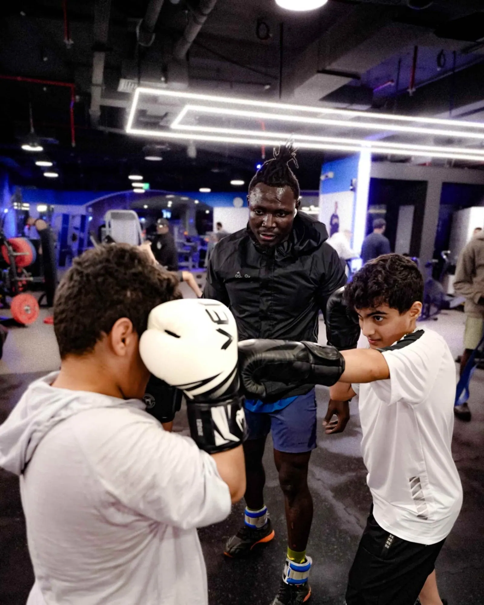 An adult guiding two children practising with boxing gloves inside a gym.