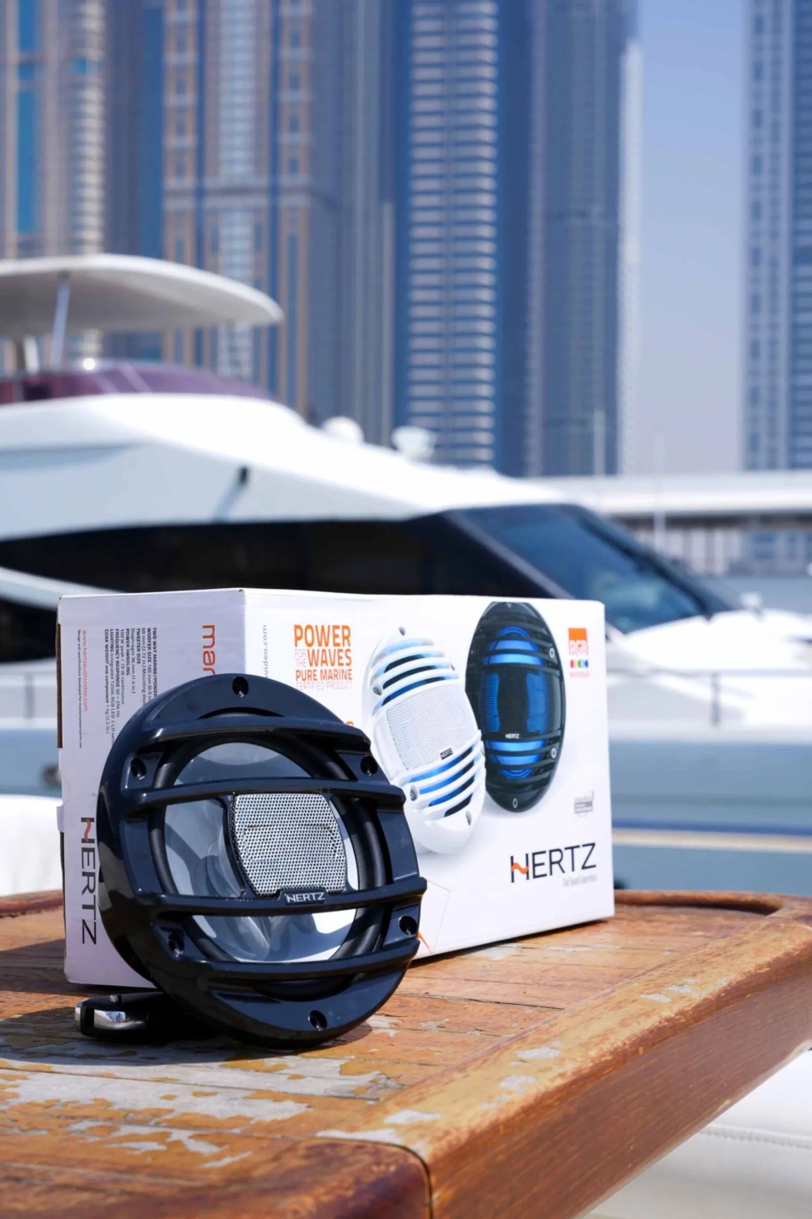 A marine speaker and its box photographed on a wooden dock, with boats and towers behind.