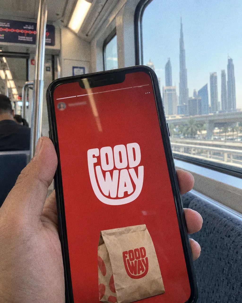 A hand holding a phone showing a red Food Way story frame with the white logo and a kraft bag, on a Dubai metro seat with a blurred window behind.