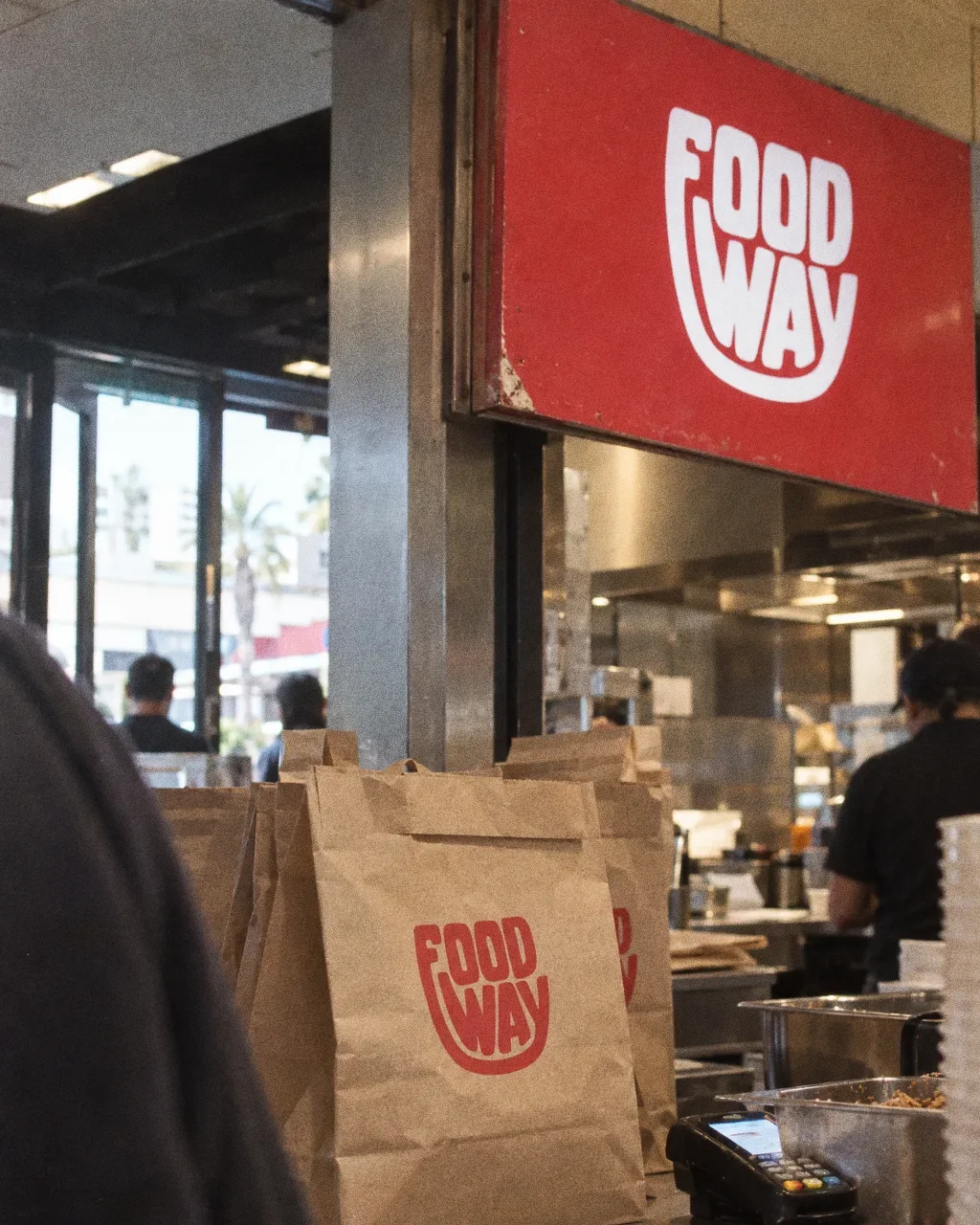 A red wall panel with the white Food Way logo above a worn order counter with kraft bags and a card machine, seen from the queue at an angle.