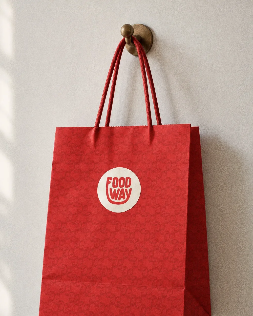 A red paper bag printed with the tonal Food Way pattern and a round cream logo sticker, hanging from a brass hook on a clean plaster wall.