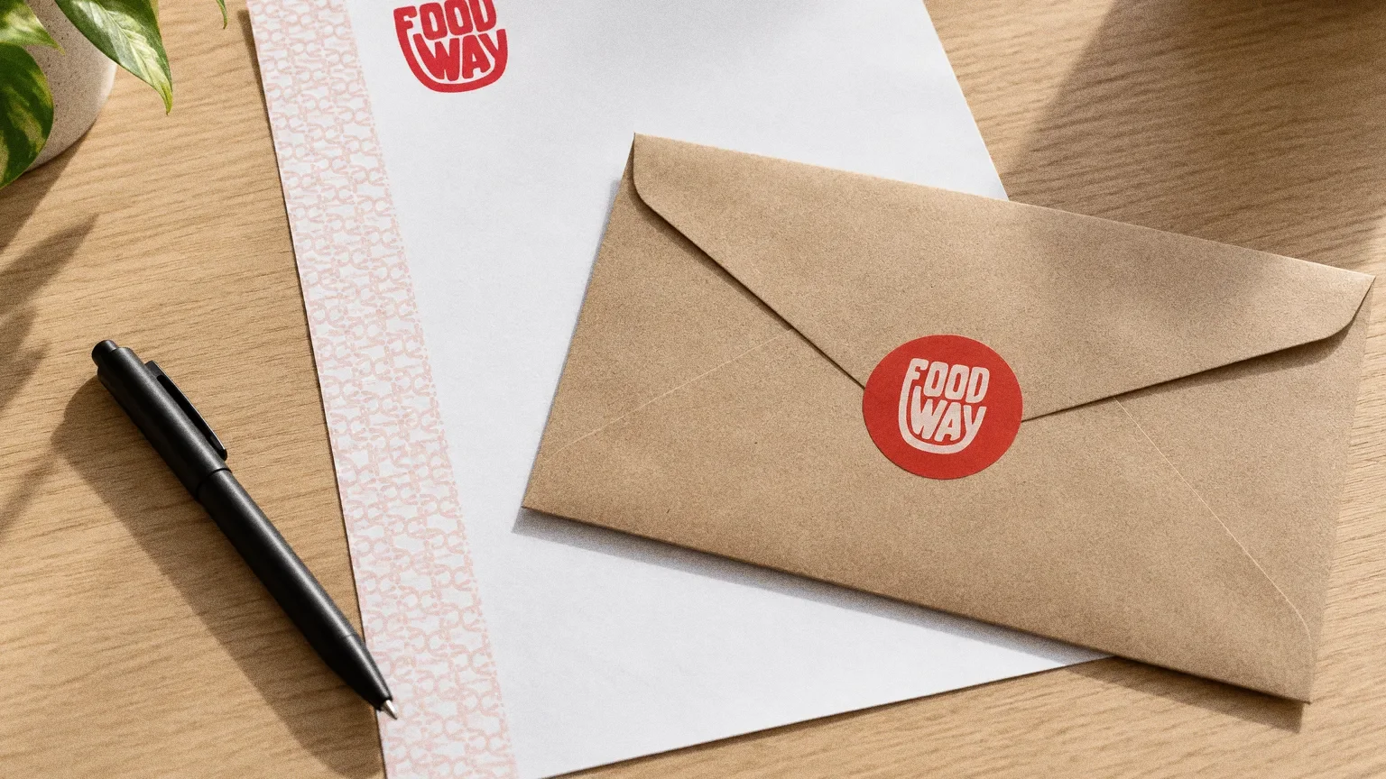 A kraft envelope sealed with a round red Food Way sticker on a white letterhead with a pink pattern edge, beside a pen on a clean oak desk.