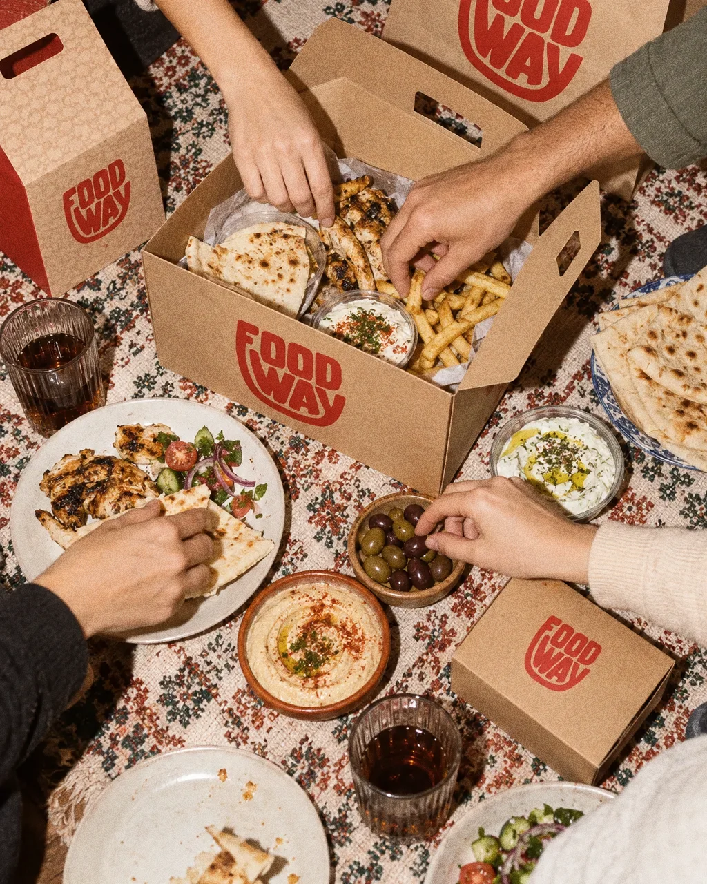 A top-down view of hands reaching into an open kraft Food Way carry box on a tablecloth among plates and dips.