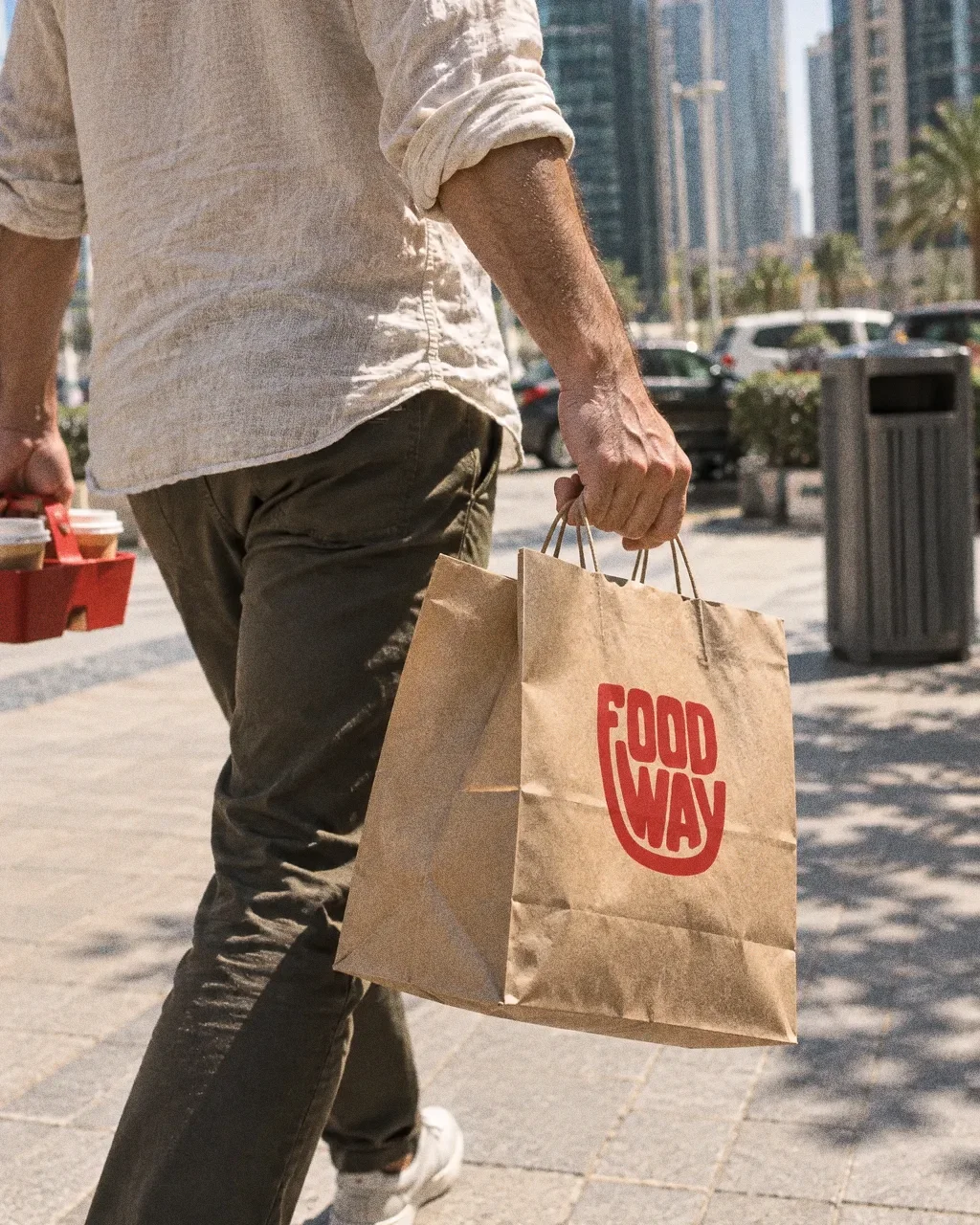 A person in a linen shirt carries a large kraft Food Way bag and a red drinks carrier along a sunny Dubai walkway with towers behind.