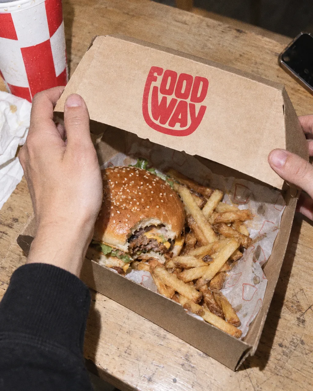 Hands lift the kraft lid of a Food Way box with the red wordmark, showing a burger and fries on patterned paper, with a red cup and a napkin beside it.