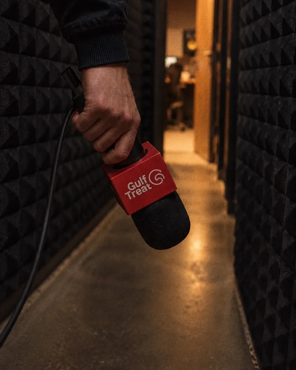 A hand carrying a black microphone with a red Gulf Treat cube flag down a dark studio corridor with acoustic foam walls.