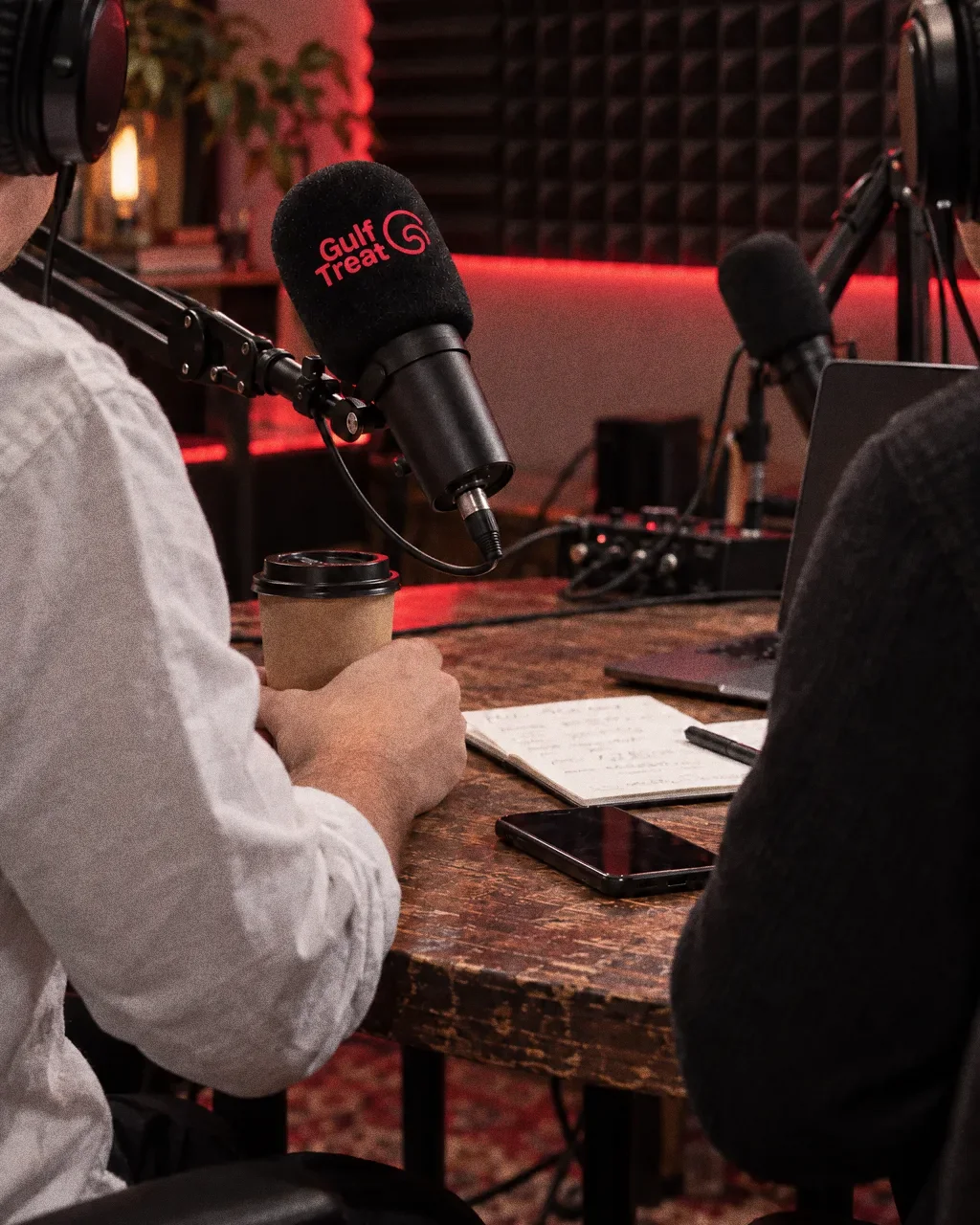 Two hosts in headphones, seen from behind, sit at a round table with boom-arm microphones carrying red Gulf Treat cube flags and windscreens, in front of a black acoustic foam wall with a red light strip.