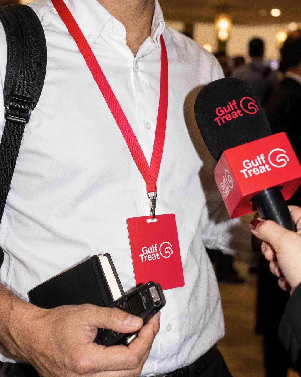 A person in a white shirt wears a red Gulf Treat lanyard and badge and holds a notebook and recorder, while a hand from the right offers a Gulf Treat microphone.