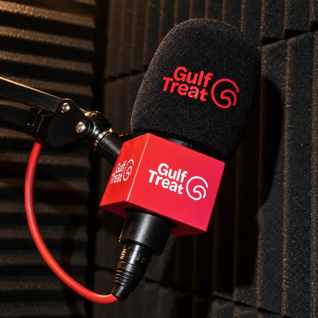 A boom-arm microphone with a black Gulf Treat windscreen and a red cube flag against black acoustic foam, with a red cable.