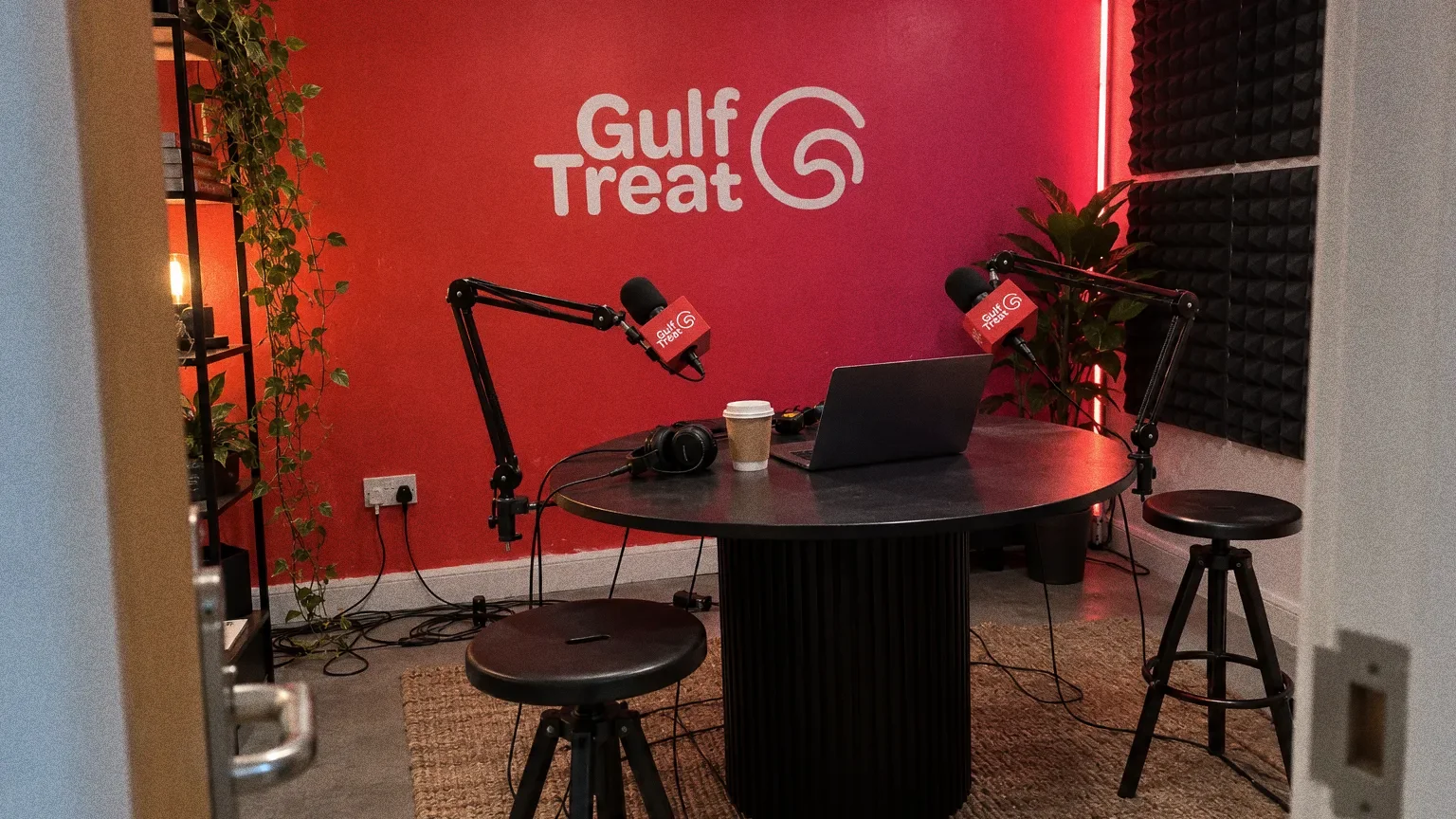 A dark broadcast studio with a red-to-pink wall carrying the white Gulf Treat wordmark and swirl G symbol, a round black desk with two microphones and two empty stools.