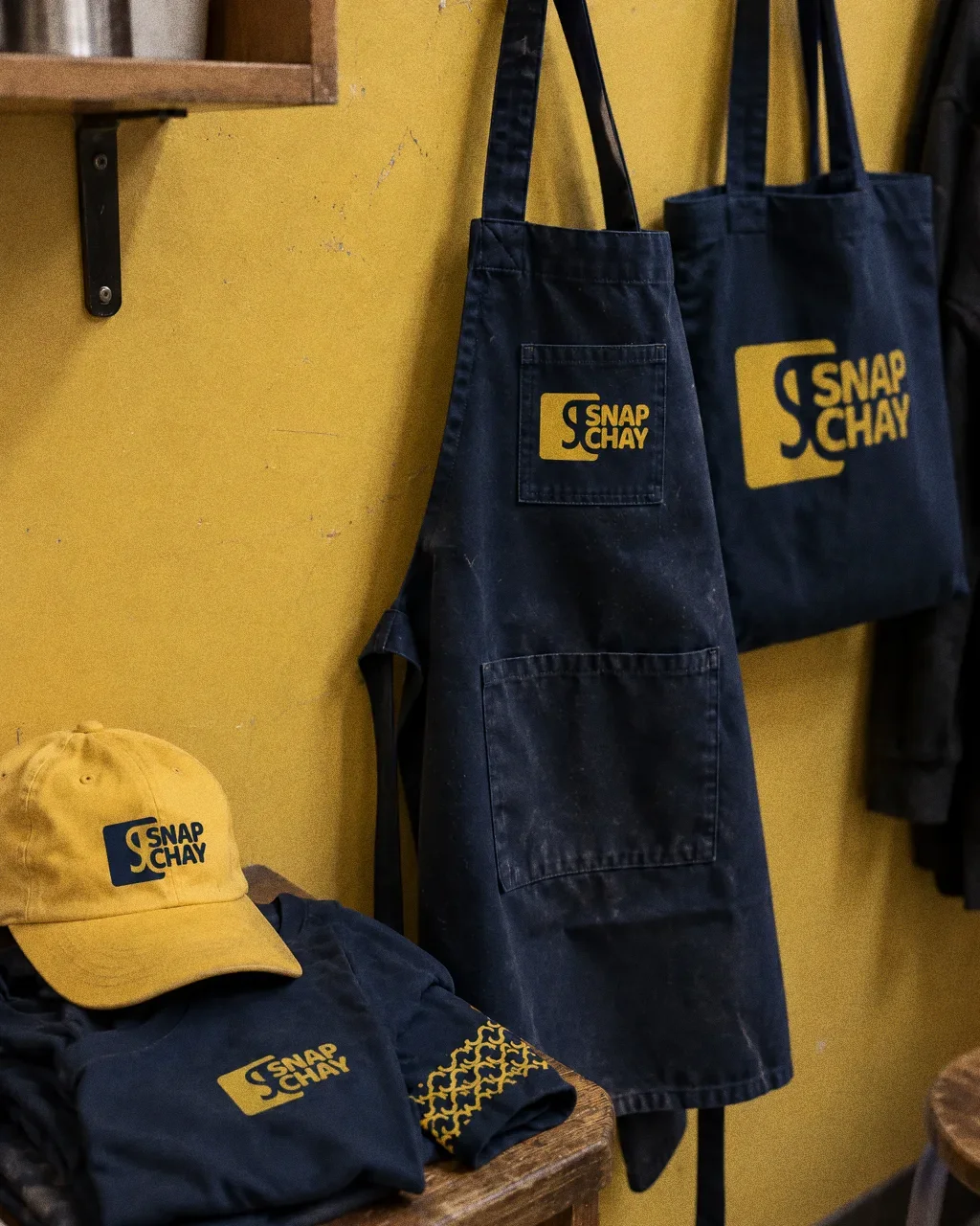 A navy apron with a yellow symbol pocket, a navy tote with a large symbol, a yellow cap and a folded navy tee, set on a yellow wall and a wooden shelf.