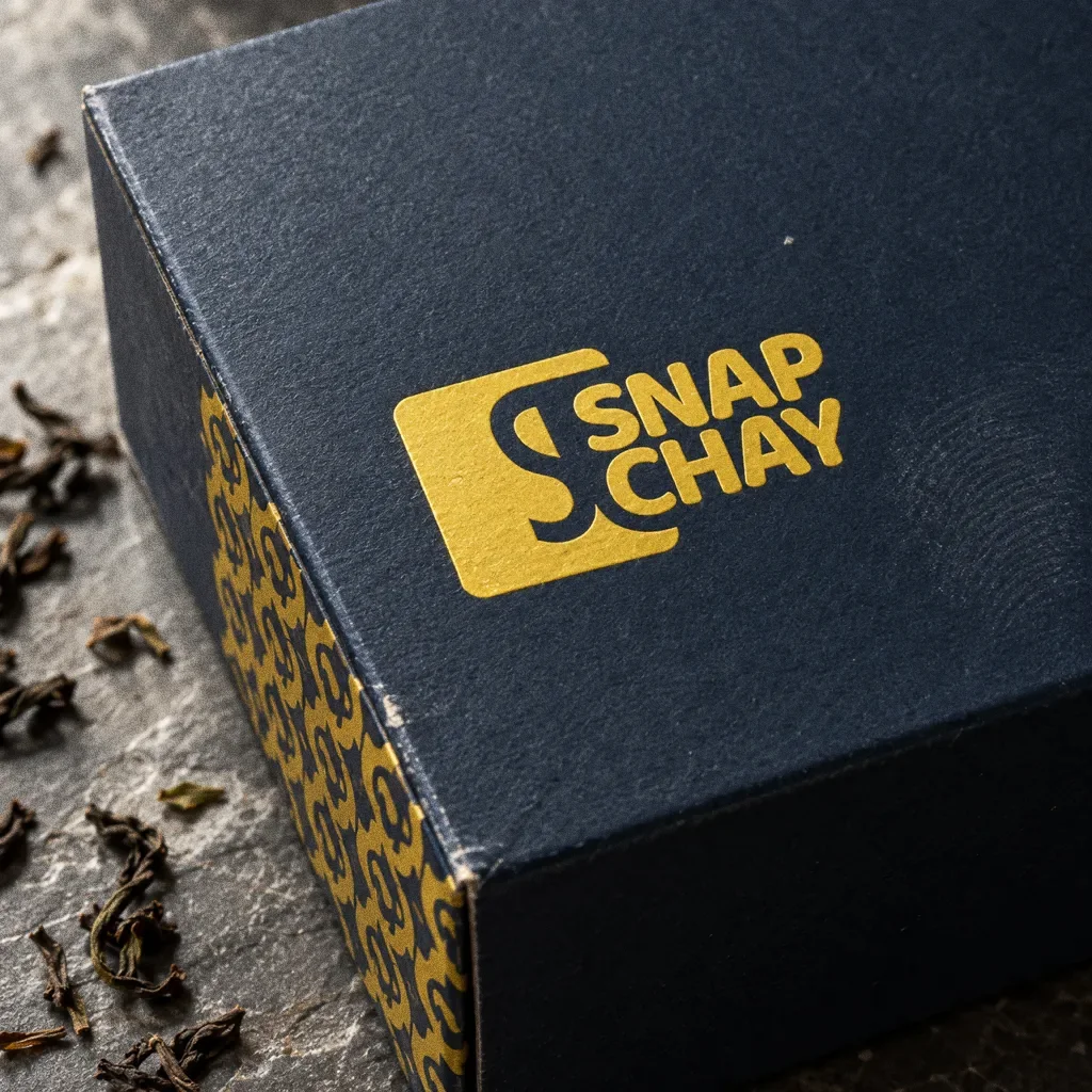 A close view of a navy tea box lid with the yellow Snap Chay tile logo and wordmark foil stamped on it, and the yellow S pattern wrapping around the corner.