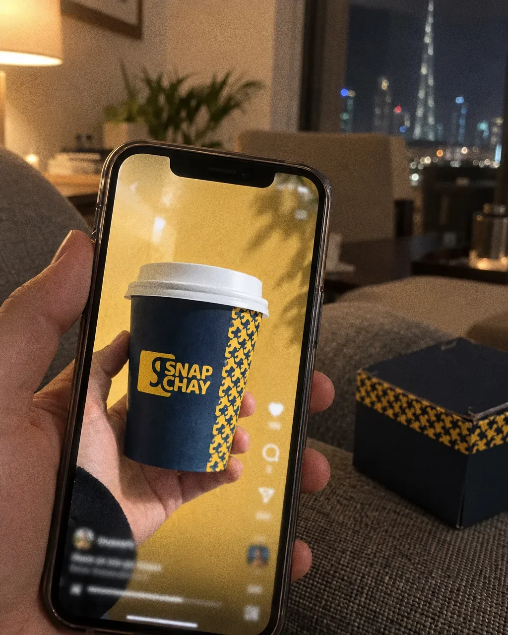 A phone held over a sofa arm showing a Snap Chay reel frame with a navy cup of chai and the yellow logo, a corner of a navy and yellow takeaway box beside it.