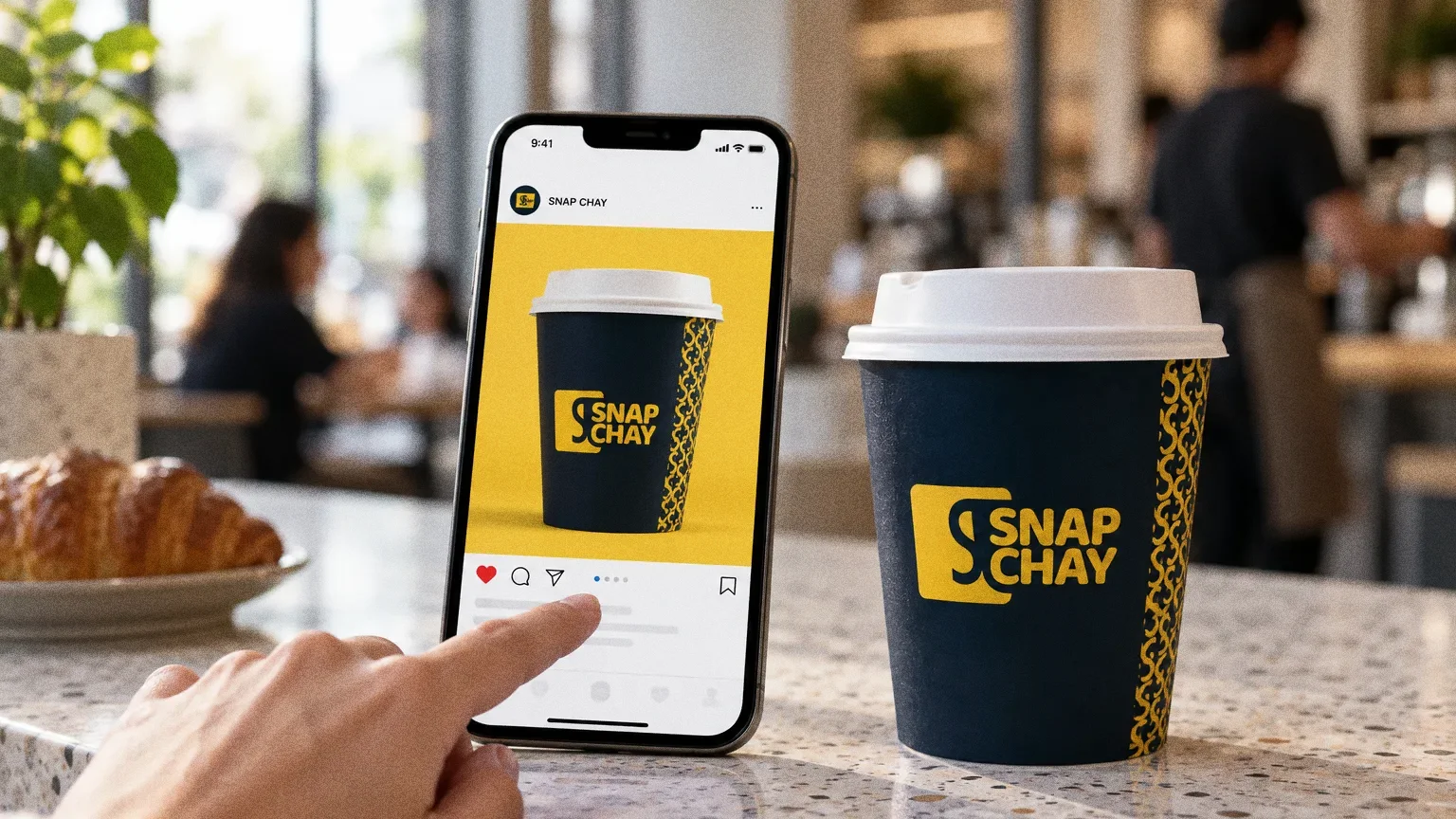 A phone on a pale terrazzo cafe counter showing a single Snap Chay post of a navy cup on yellow, with a finger touching the screen and a navy Snap Chay cup beside it.