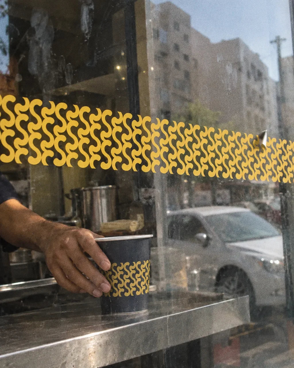A glass takeaway window with a band of the yellow interlocking Snap Chay pattern as a decal, fingerprints on the glass and a hand reaching a cup.