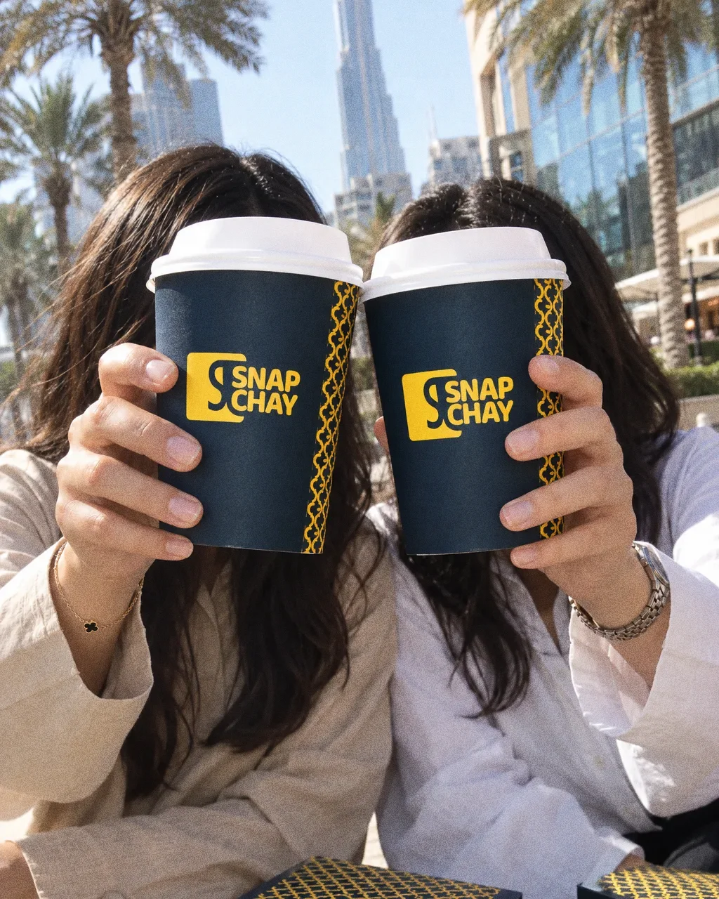 Two friends on a sunlit stone wall, each holding a navy Snap Chay cup up in front of their face as the cups touch.