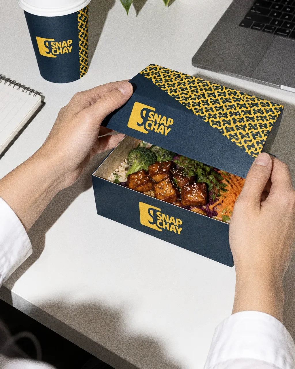 Two hands opening a navy Snap Chay takeaway box with a yellow pattern on an office desk, with a navy cup nearby.