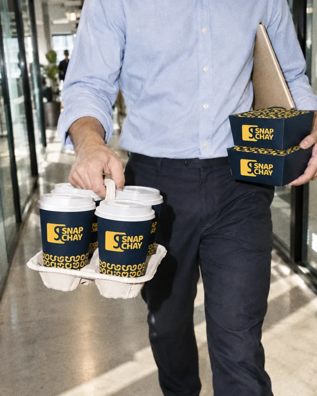 A person seen from the chest down in an office corridor carrying a carrier of navy Snap Chay cups and a folder.