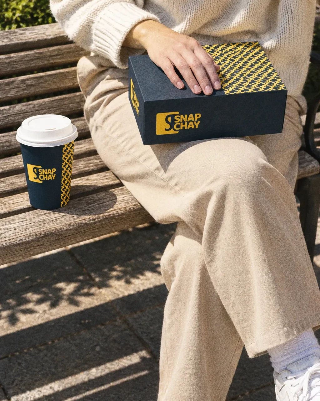 A person seen from the waist down on a sunlit bench with a navy Snap Chay box on the lap and a cup beside them.