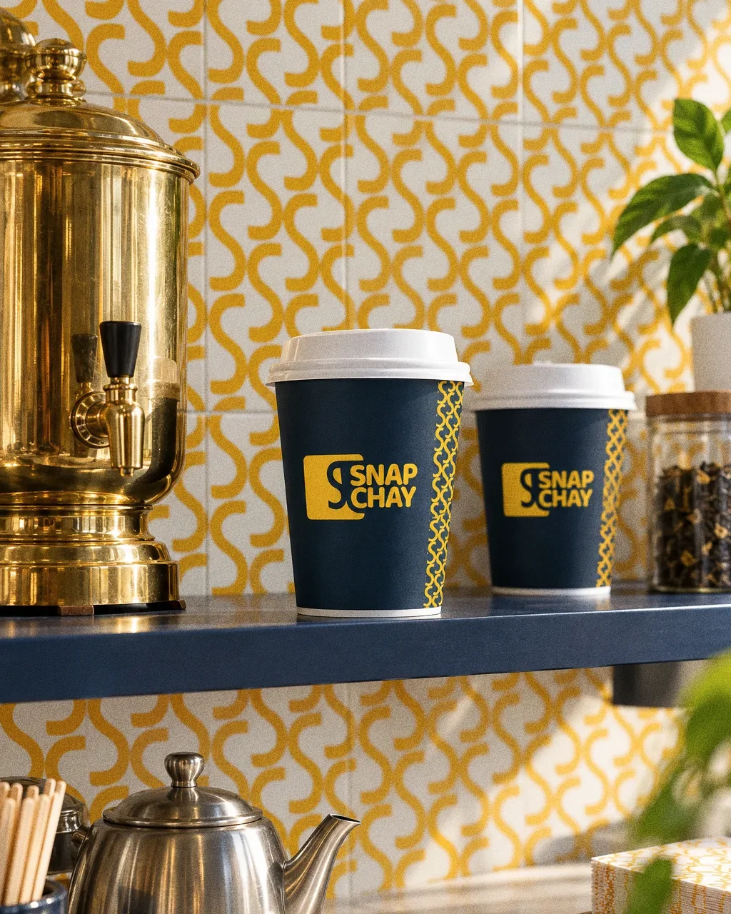 A tea counter with a yellow S-pattern tiled wall, navy Snap Chay cups on a navy shelf, a polished brass urn and a steel teapot in a bright, tidy shop.