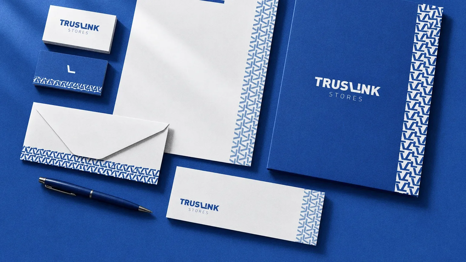 A flat-lay on blue paper of Truslink Stores business cards, a letterhead with a zigzag pattern edge, an envelope, a compliment slip, a pen and a blue folder.