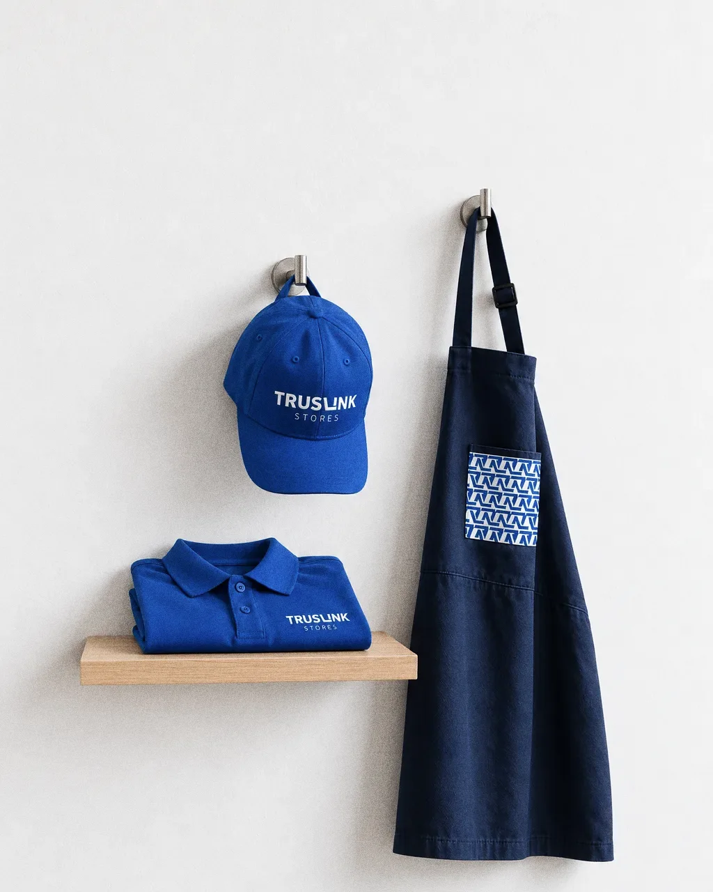 A royal blue staff cap, a navy apron with a zigzag pocket and a folded blue polo on a small shelf, hung on a clean white wall.