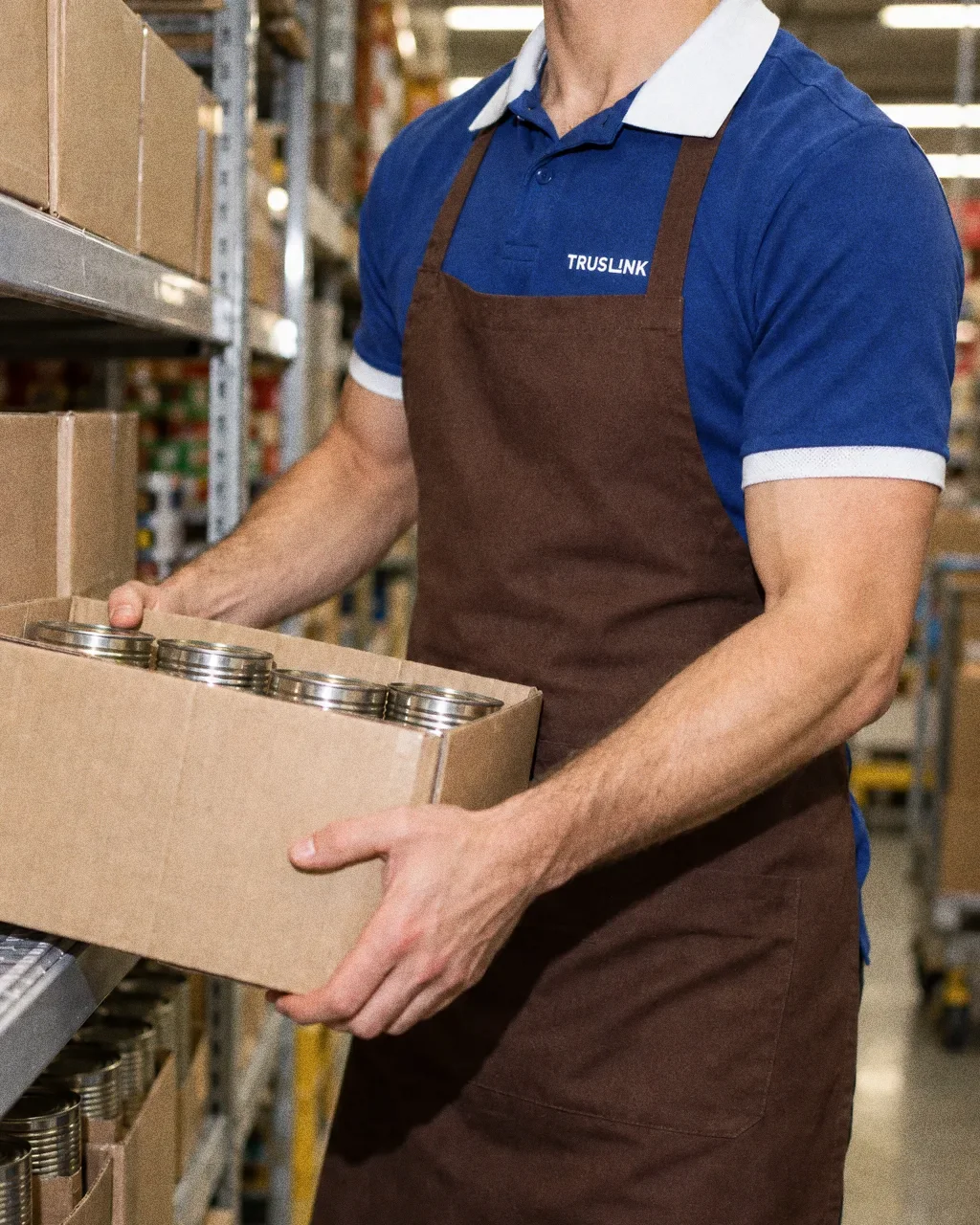 A Truslink staff member in a blue polo with white collar and a brown apron, seen from the chest down, lifting a carton of tins onto a stockroom shelf.