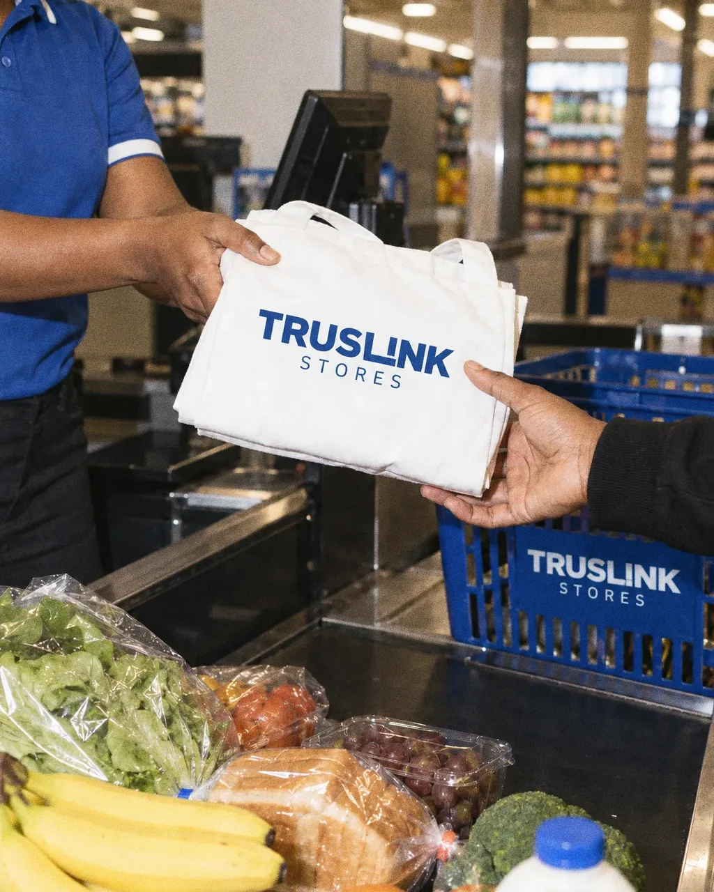 A cashier in a blue polo passes a white Truslink Stores tote bag to a customer at the till, with groceries and a blue Truslink basket on the counter.